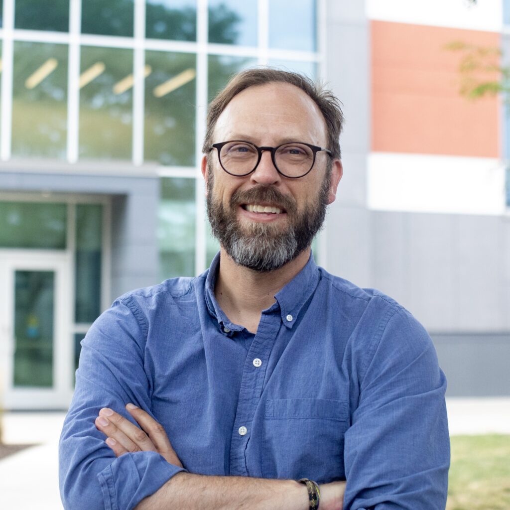 headshot of Brock Kirwan wearing a blue button-down shirt with his arms crossed standing in front of an office building. He is bespectacled has short brown hair and a graying beard which he thinks is handsome though his family have differing opinions on this point. He is standing in the sunshine and looks happy.